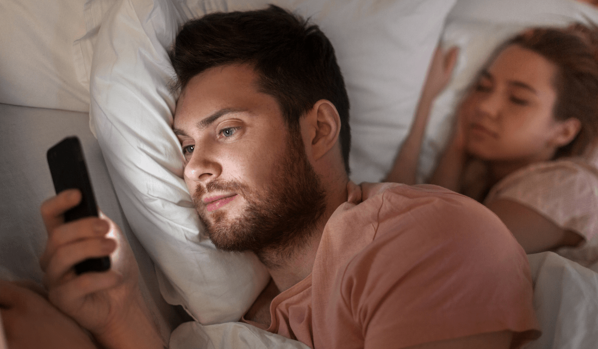 couple in bed and man looking at his mobile messages while woman sleeps