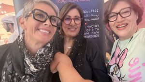 A close-up selfie of three women smiling warmly at the premiere of Shades of That Light. Sahar stands on the right. The director, Nujoom Al Ghanem, and the artist stand beside her in front of the film’s promotional banner. The setting is bright and lively, with people in traditional Emirati attire visible in the background.