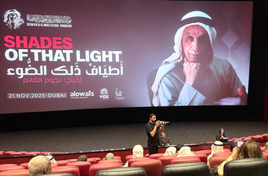 A wide-angle photograph of the cinema hall at VOX Wafi Mall during the premiere of Shades of That Light. The large screen displays the film’s title in English and Arabic, along with the portrait of Sultan Bin Ali Al Owais. The audience sits in red theatre seats, while a photographer stands near the front capturing the event. The atmosphere is attentive and formal, with Emirati attendees dressed in traditional clothing.